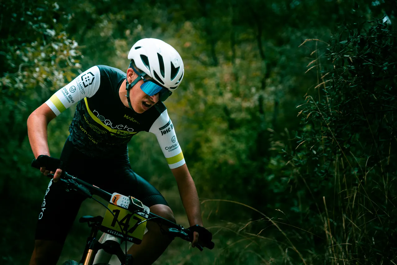 Ciclista amb casc blanc avançant entre vegetació.